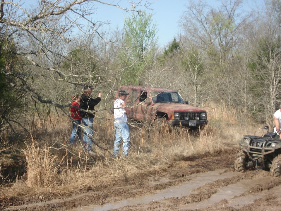 Rusty Jeep Cherokee XJ stuck in mud with people around it and an ATV nearby