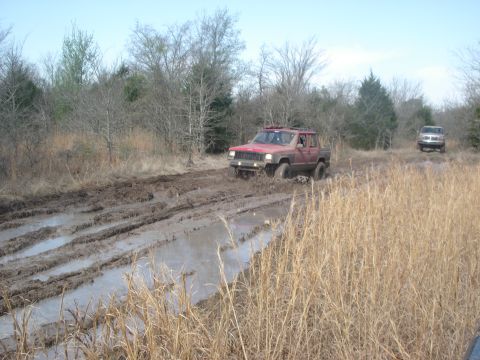1989 Ford Bronco II - Two SUVs navigate a muddy off-road trail with dry grass and trees in the background.