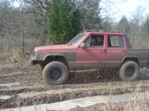 1989 Ford Bronco II - A red Jeep Comanche pickup truck navigates a muddy trail, kicking up dirt as it drives.