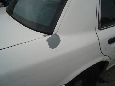 2000 Ford Crown Victoria - The rear quarter panel of a white sedan shows significant paint damage, with primer exposed in a large circular area.