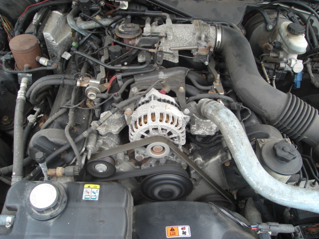 Engine bay with alternator, belts, and hoses visible. Some corrosion on metal parts