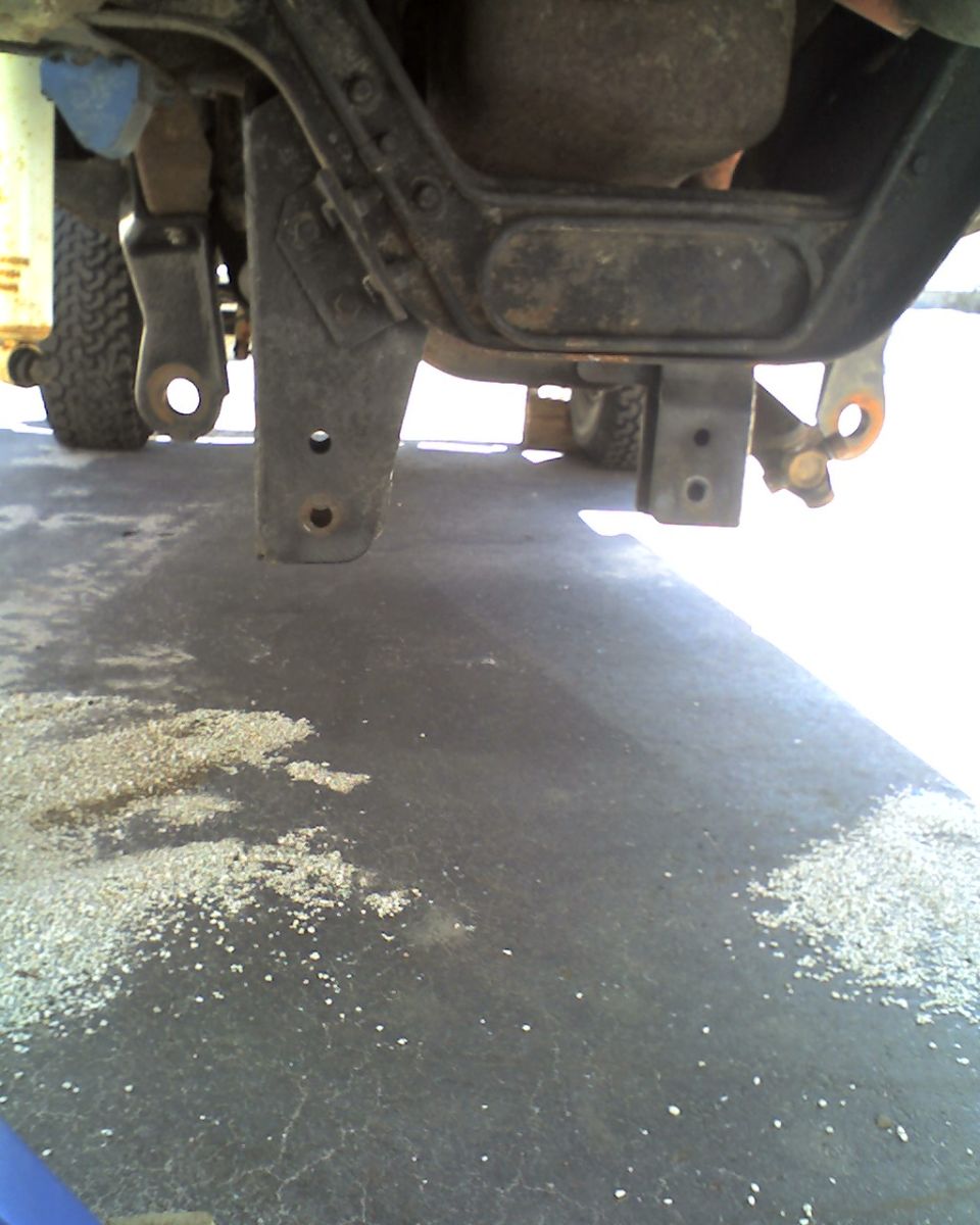 Undercarriage of a truck with visible suspension components and a filter. The ground is asphalt with scattered debris
