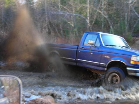 1996 Ford F-150 - A blue 1996 Ford F-150 navigates a muddy, watery obstacle, creating a significant splash.