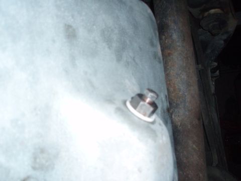 1987 Ford Bronco - Close-up of an oil pan drain plug, a white gasket and a hex head, with a rusty pipe in the background.