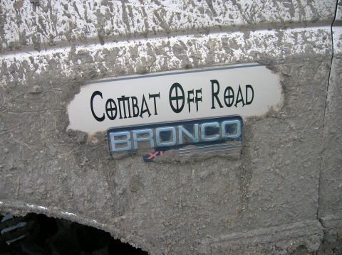 1996 Ford Bronco - Mud-splattered white body panel with a chrome 'BRONCO XL' emblem below a white decal reading 'COMBAT OFF ROAD.' Thick brown mud coats the s…