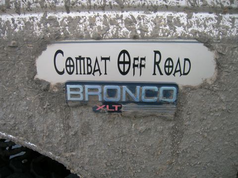 1996 Ford Bronco - Close-up of a mud-caked white body panel bearing a chrome 'BRONCO XLT' emblem and a 'COMBAT OFF ROAD' decal above it. Thick dried mud coats…