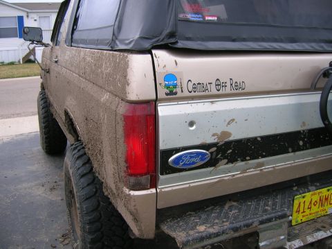 1996 Ford Bronco - Mud-caked rear quarter of a lifted tan and silver two-tone Ford SUV with a black soft top, oversized mud-terrain tires and a swing-away tir…