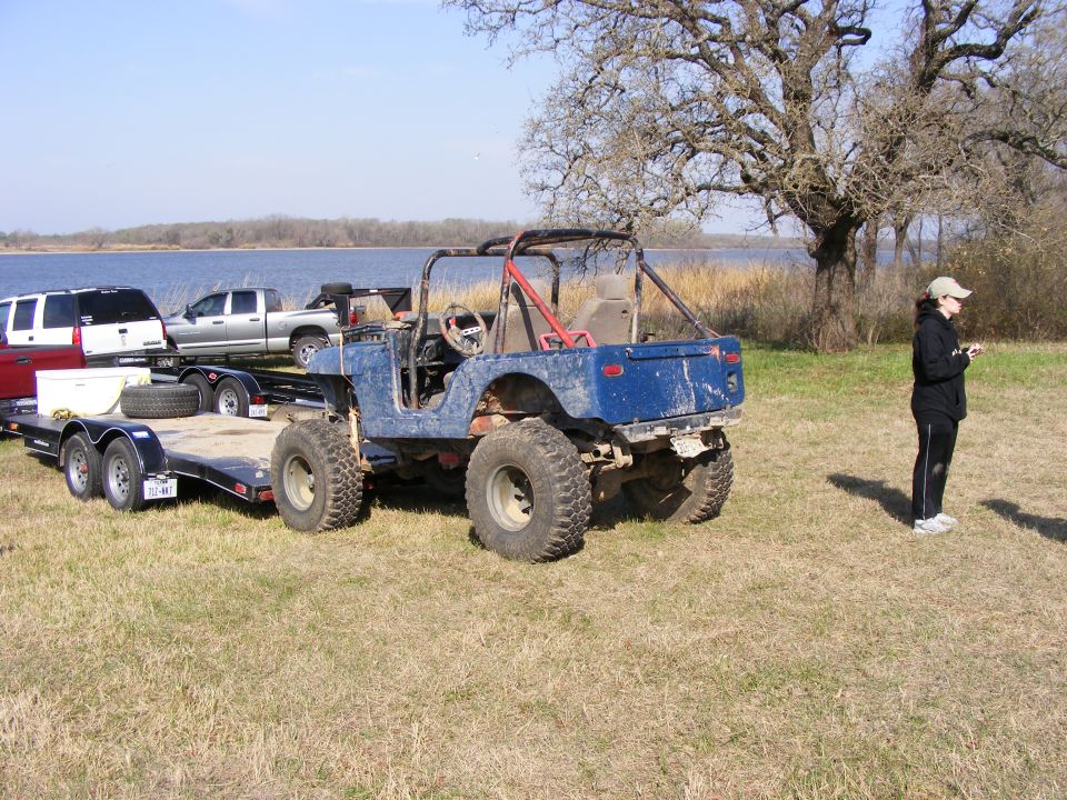 Blue Jeep with roll cage parked next to a trailer with a white SUV and a pickup truck. Person stands nearby