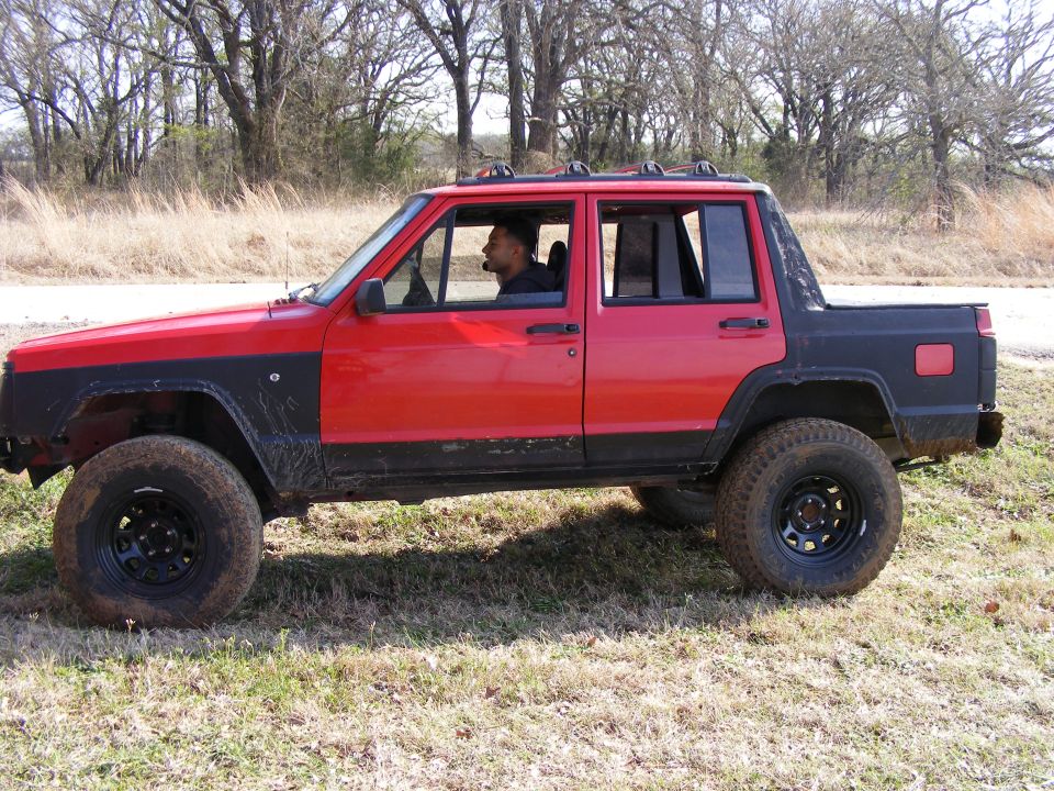 Red and black Jeep Cherokee pickup truck with black fender flares and roof rack, parked on grassy terrain