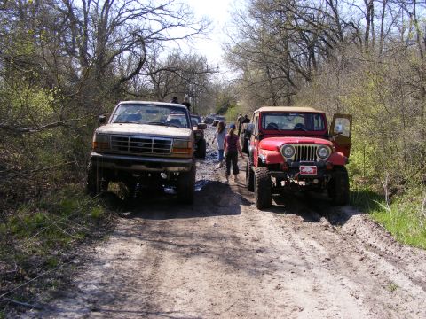 1981 Jeep CJ7 - Two off-road vehicles, a red Jeep CJ7 and a lifted Ford truck, are stopped on a muddy path surrounded by trees.