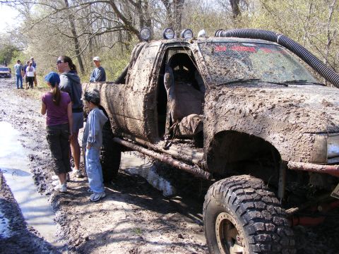 1981 Jeep CJ7 - Off-road enthusiasts gather around a heavily mud-caked pickup truck on a challenging trail.