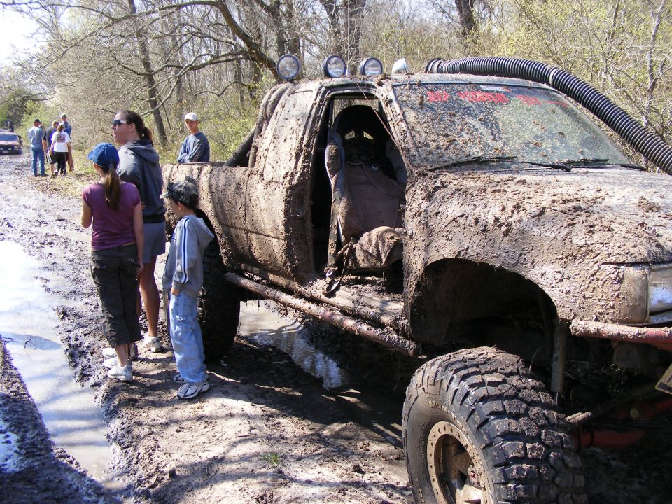 Mud-covered pickup truck with snorkel and roll cage, parked on a muddy trail with people watching. Another vehicle is visible in the background