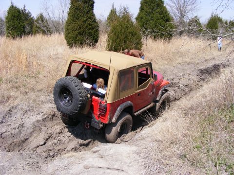 1981 Jeep CJ7 - A red Jeep CJ7 tackles a challenging muddy off-road course, with passengers inside and a spare tire mounted on the back.