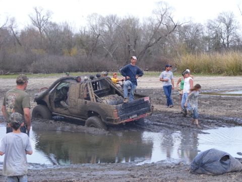1981 Jeep CJ7 - A group of people observe a heavily modified pickup truck stuck deep in a muddy water hole during an off-road event.