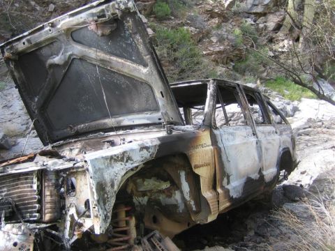 1987 Suzuki Samurai - A heavily damaged SUV sits in a rocky, outdoor environment, its hood ajar and exterior charred.