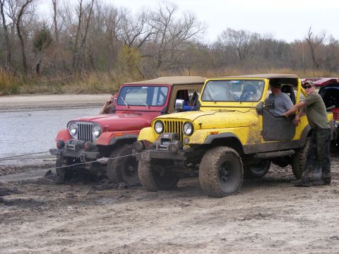 1981 Jeep CJ7 - Two Jeep CJ series vehicles are stuck in the mud next to a body of water, with people interacting with them.