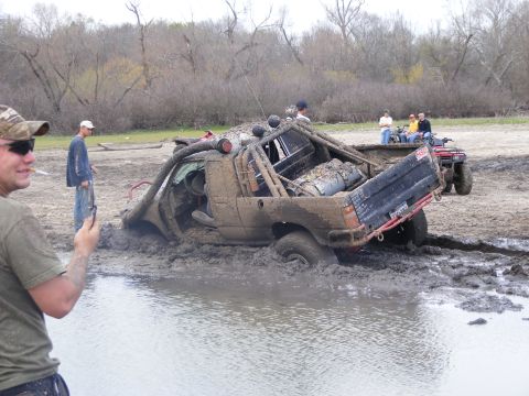 1981 Jeep CJ7 - An off-road vehicle is deeply stuck in mud and water, with onlookers and an ATV present at the scene.