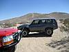 1986 Ford Bronco - A dark SUV is parked on a dusty road in a desert landscape, with a red vehicle partially visible.