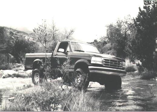 Dark Ford F-150 regular cab pickup fording a shallow creek among trees, black-and-white photo