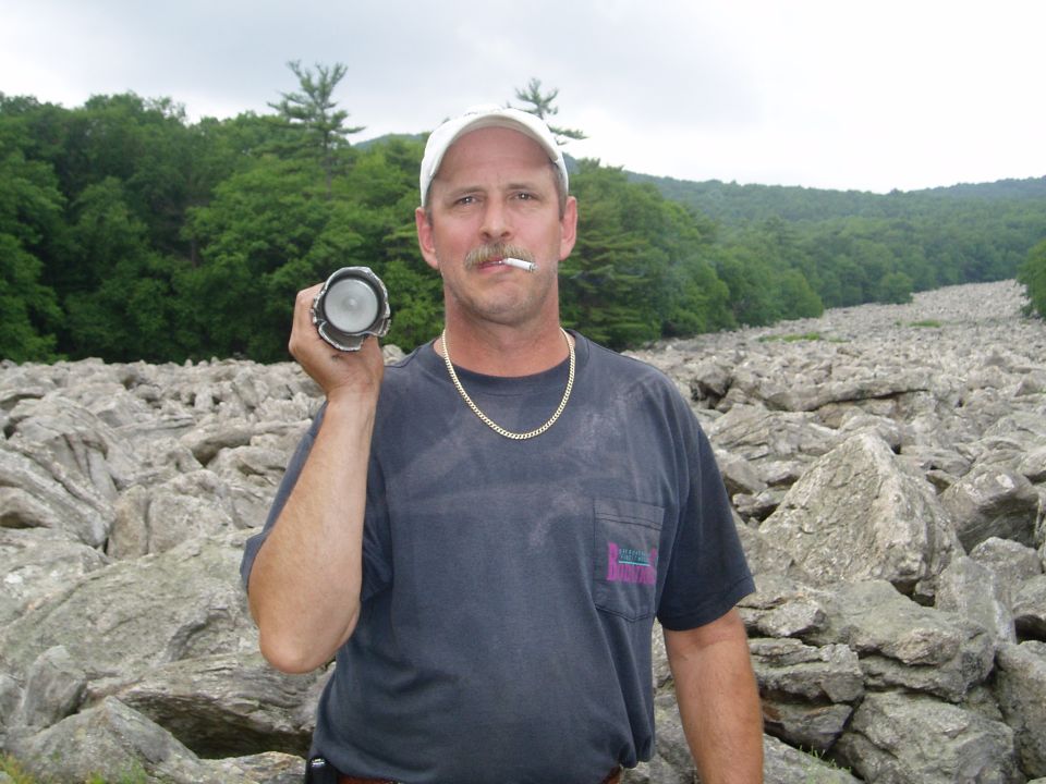 Man in a dark t-shirt and white cap holds a flashlight, smoking a cigarette, with a rocky landscape behind him
