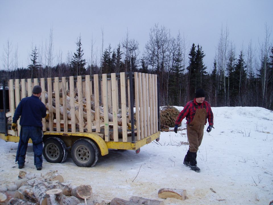 Two men in a snowy, wooded area with a trailer full of cut firewood