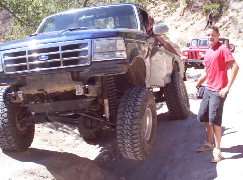 1992 Ford F-150 - A lifted 1992 Ford F-150 with oversized tires is shown off-road, with another vehicle visible in the background.