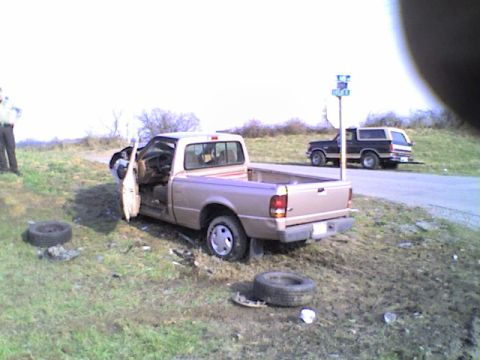 1990 Ford Bronco - A tan Ford Ranger pickup truck sits off the road with its driver door open and a tire removed, while a black Ford Bronco is parked on the r…
