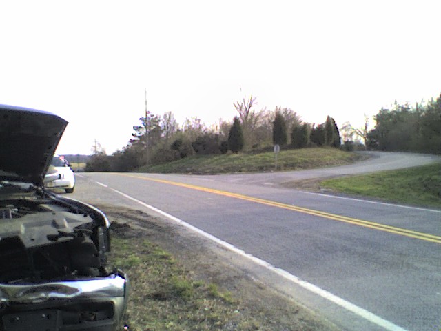 Dark vehicle front with hood open, white car in background, roadside
