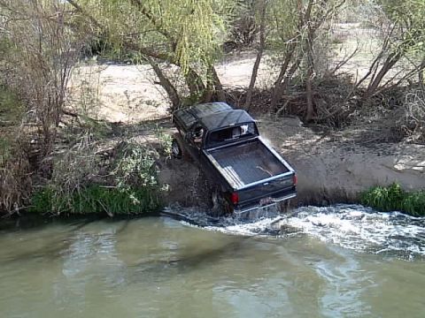 1992 Ford F-150 - A black pickup truck navigates a shallow body of water near a wooded bank.