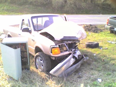 1990 Ford Bronco - A heavily damaged gold pickup truck sits on the side of the road next to a utility box, with another vehicle visible in the background.