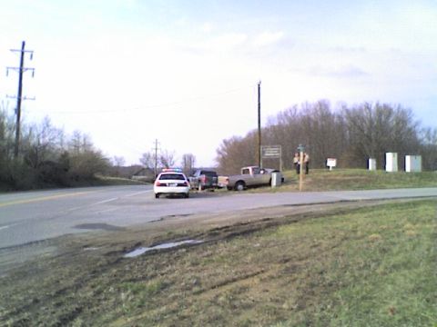 1990 Ford Bronco - A police car and a pickup truck are stopped near a road with trees in the background.