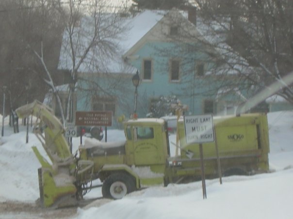 Yellow snow blower truck parked in snow, with a light blue house in the background. Signs indicate traffic directions