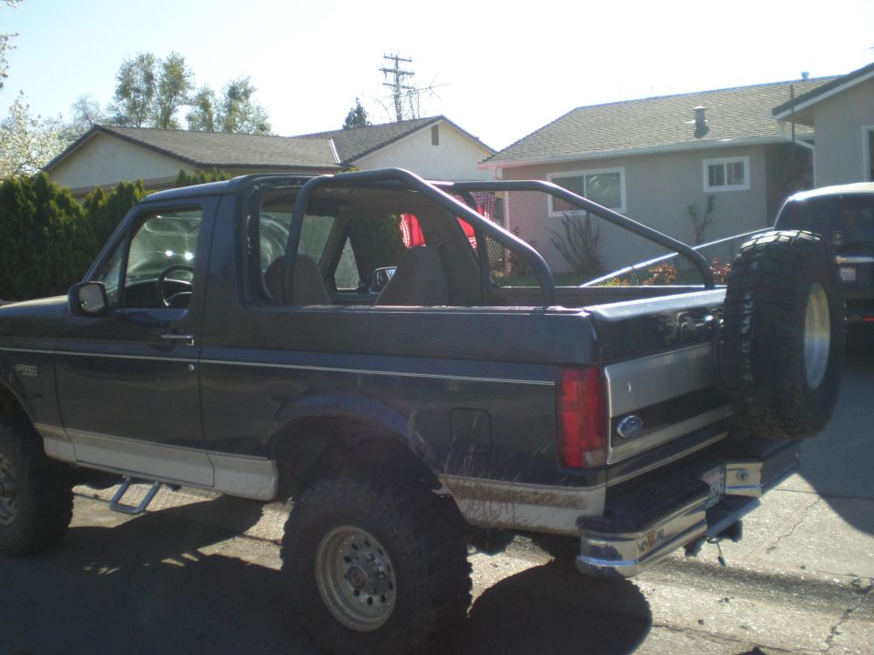 Rear side view of lifted dark blue Ford Bronco with top removed, roll bar and spare tire