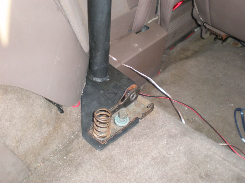 Black shifter base on rusty bracket with coil spring over tan carpet, loose wiring nearby