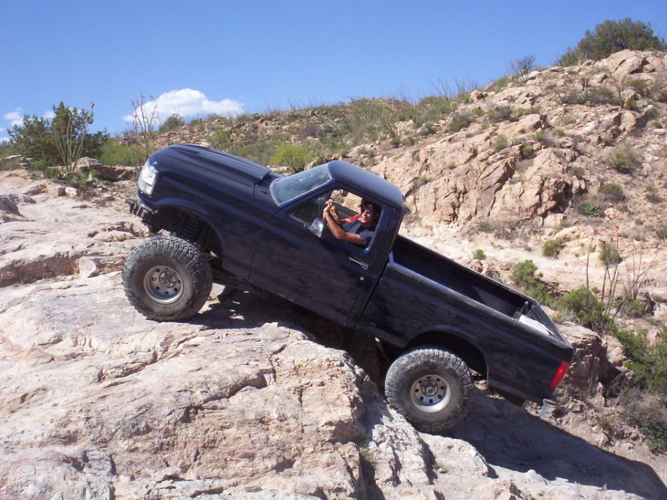 Dark blue lifted Ford pickup on oversized tires climbing a steep rock ledge in desert terrain