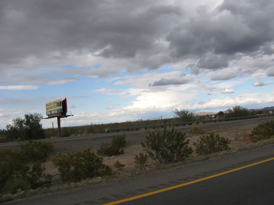 Billboard with colorful advertisements stands beside a highway under a cloudy sky