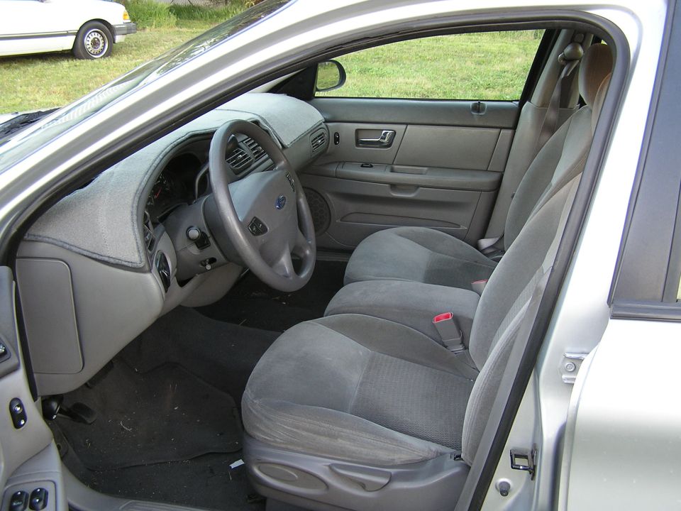 Gray cloth interior of a silver Ford sedan seen through the open driver's door, with carpeted dash cover