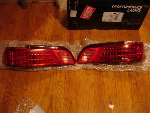 1993 Ford Taurus - Two red LED tail lights are displayed on a wooden floor next to a box of performance lamps.