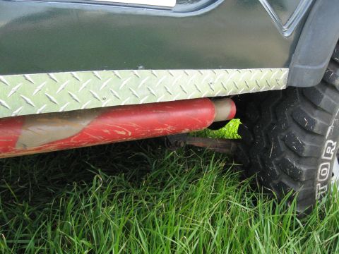 1988 Ford Bronco - Close-up of the side of a green Jeep shows diamond plate trim and a red exhaust pipe against a grassy background.