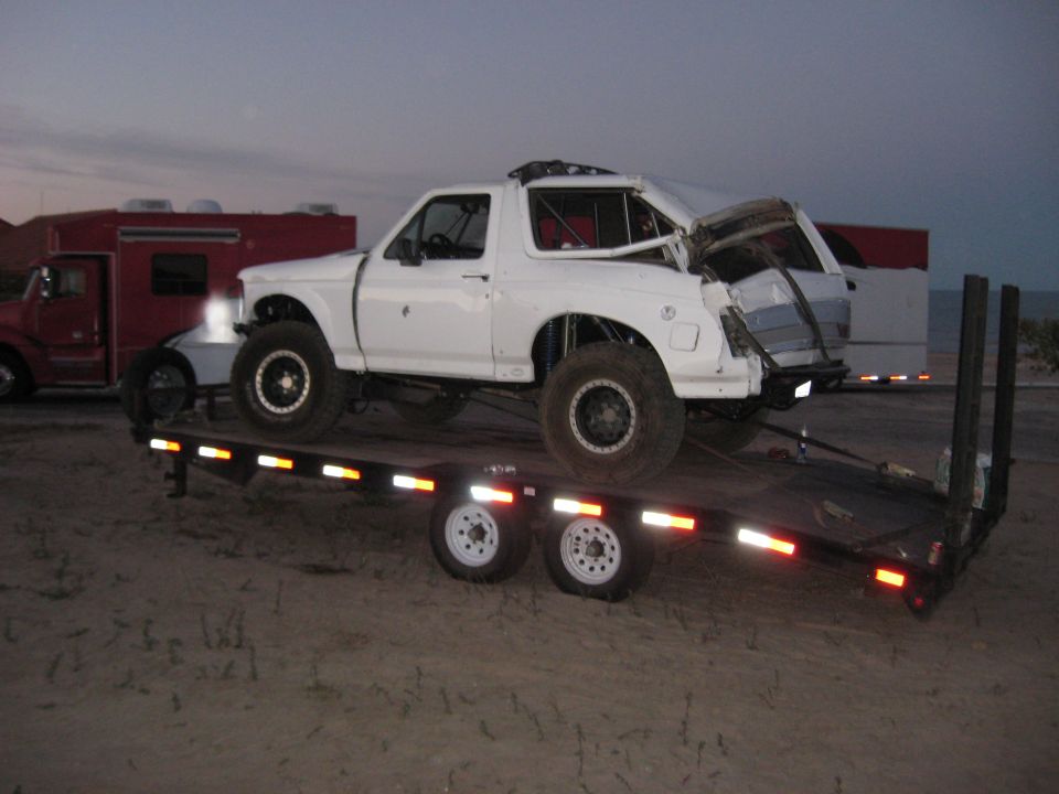 White off-road Bronco with damaged front end and roll cage on a flatbed trailer at dusk