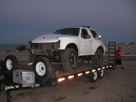 1994 Ford Bronco - A white SUV is loaded onto a trailer, its front end appears damaged. A person stands on the trailer ramp.