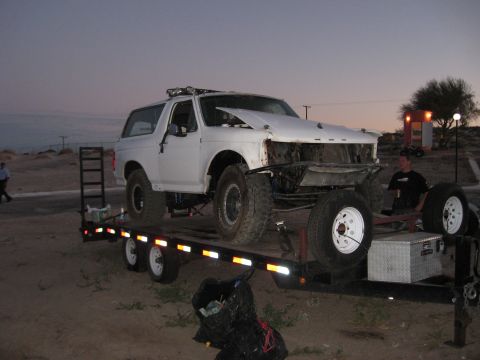 1994 Ford Bronco - A white Ford Bronco is loaded onto a trailer, showing significant damage to its front end and large off-road tires.
