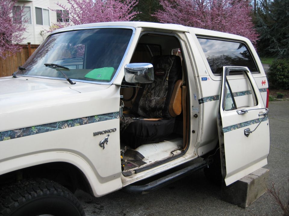 White Ford Bronco with camouflage seat covers and door removed. Pink flowering trees in background