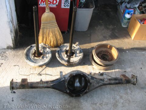 1987 Suzuki Samurai - Vehicle axle and brake parts are laid out on a concrete surface, with a broom and tool chest in the background.