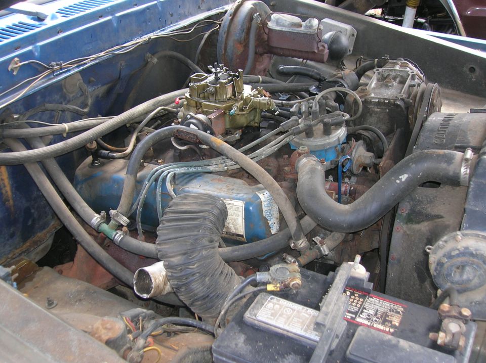 Engine bay with carburetor, distributor, hoses, and battery visible. Some text on battery and radiator shroud