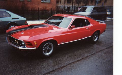 1998 Ford Crown Victoria - A red 1970 Ford Mustang coupe is parked on a wet street next to other vehicles.