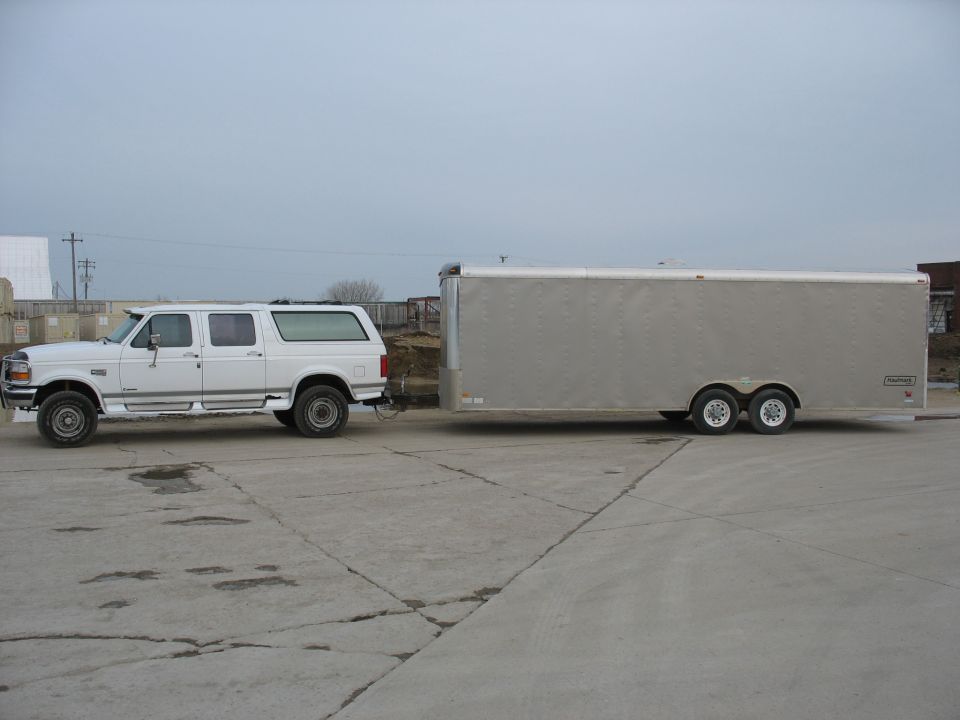 White Ford Bronco SUV towing a silver enclosed trailer. The SUV has four doors and a custom rear section