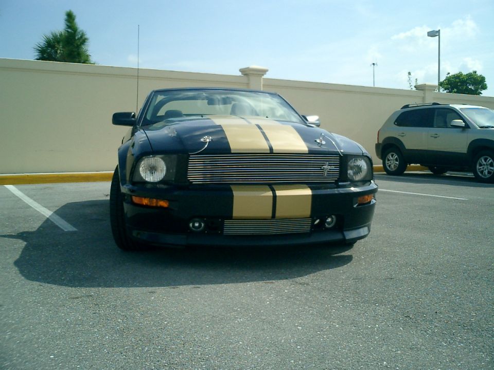 Black convertible Mustang with gold stripes parked in a lot, another SUV is behind it