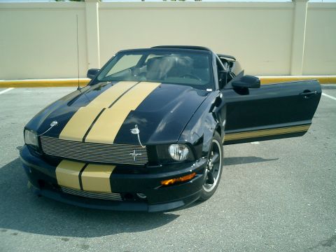 2007 Ford Mustang - Black 2007 Ford Mustang convertible with gold racing stripes and driver's door ajar, parked outdoors.