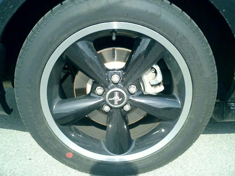 Close-up of a black 5-spoke wheel with a silver lip on a 2007 Ford Mustang. The Mustang logo is visible in the center cap